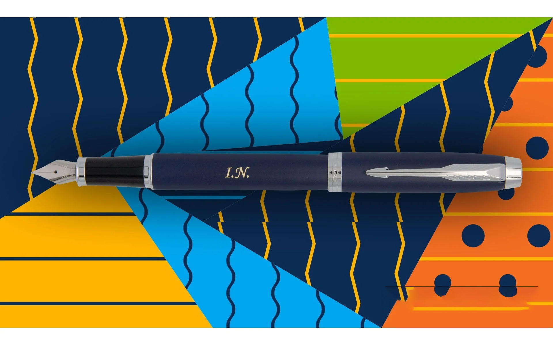 Pióro wieczne Parker IM Niebieskie CT [1931647] – eleganckie granatowe pióro z wykończeniami w kolorze chromu (CT – Chrome Trim), grawer: „I.N.”. Umieszczone na tle kolorowej, geometrycznej grafiki w odcieniach żółci, błękitu, zieleni i pomarańczu.