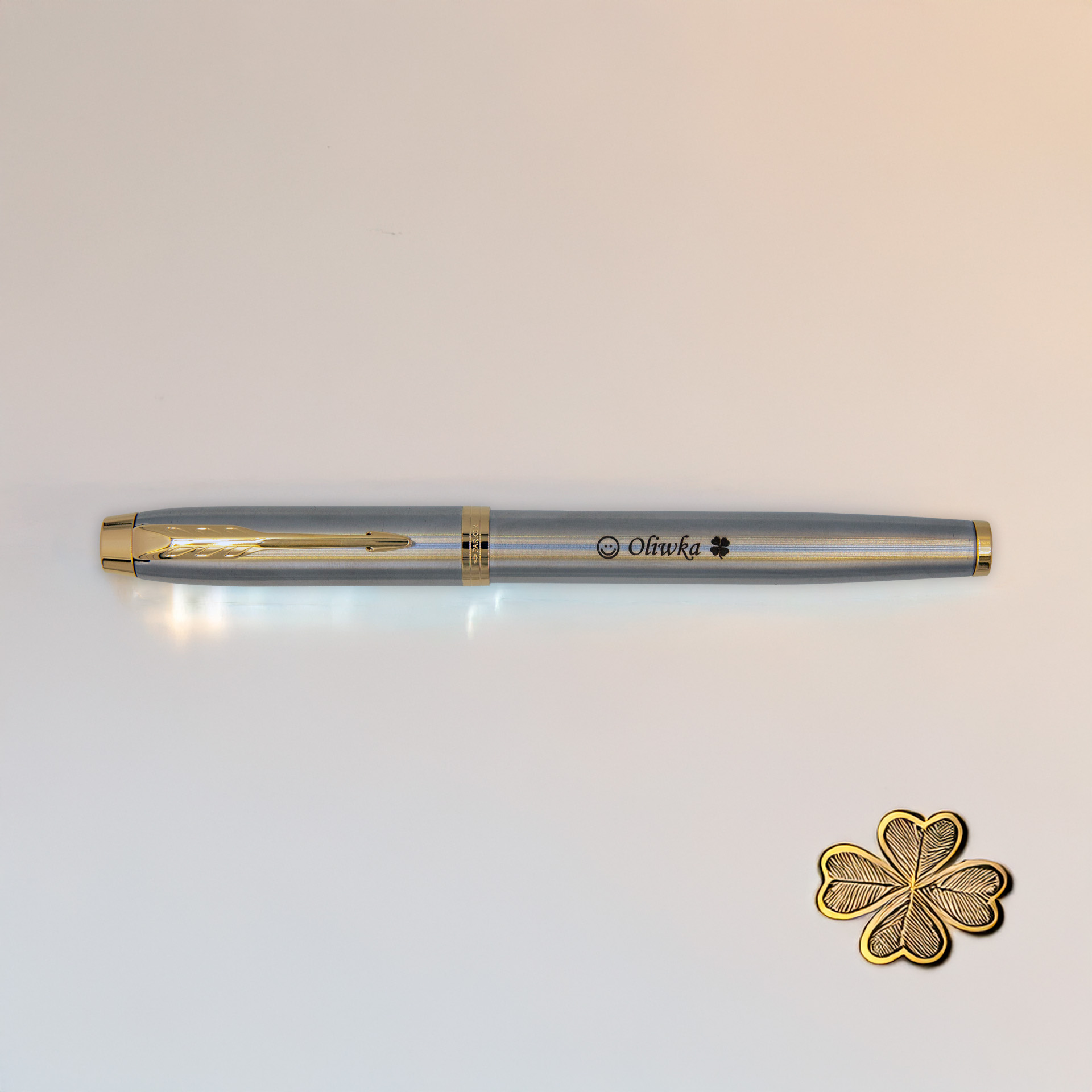 Pióro wieczne Parker IM Brushed Metal GT (1931649) z grawerem „Oliwka” i symbolem koniczynki, sfotografowany na jasnym tle obok złotej czterolistnej koniczynki.